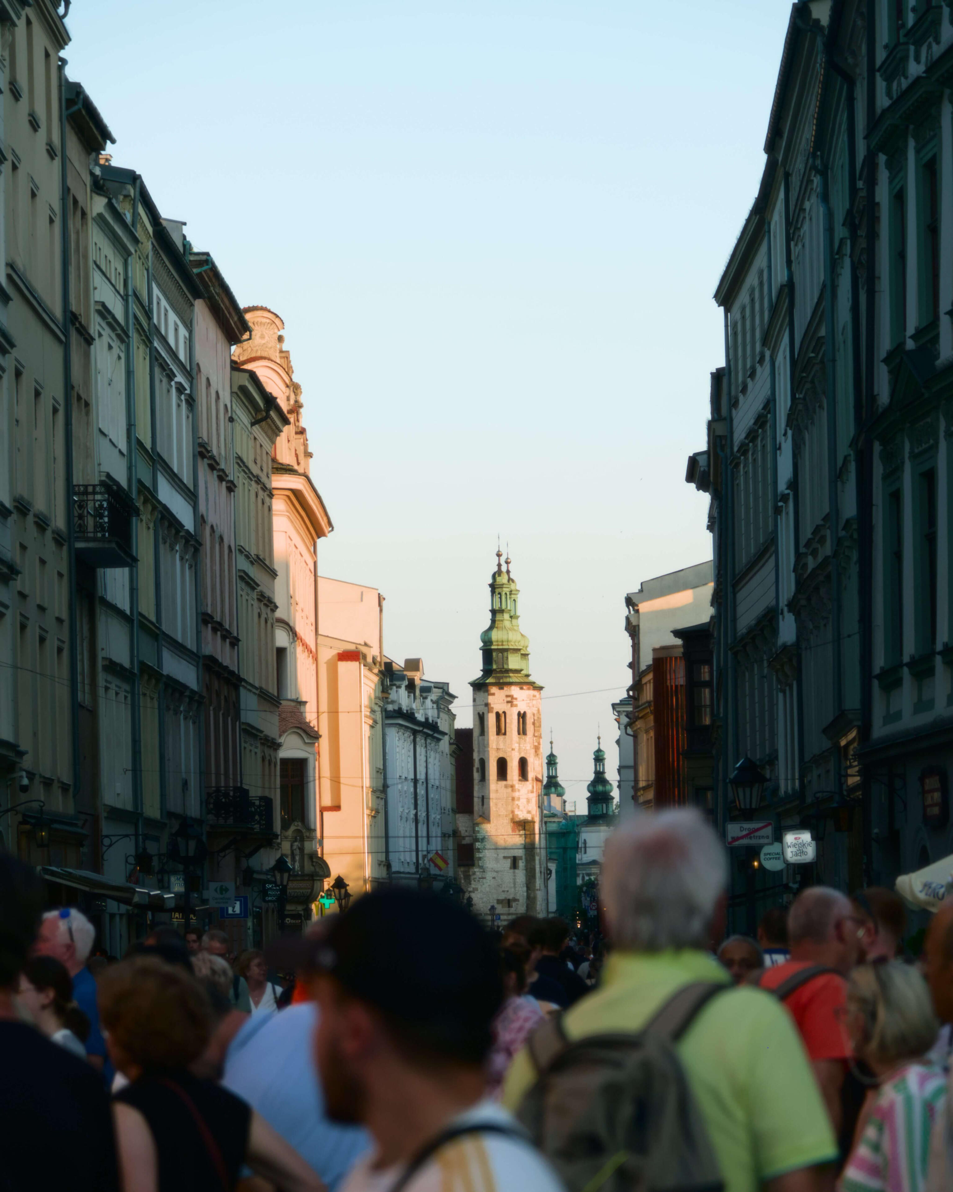 Zatłoczona ulica Grodzka w Krakowie podczas złotej godziny. W oddali widoczna są wieże kościoła Św. Andrzeja i kościoła Bernardynów. Na pierwszym planie znajduje się tłum przechodniów ustawionych tyłem i bokiem do aparatu